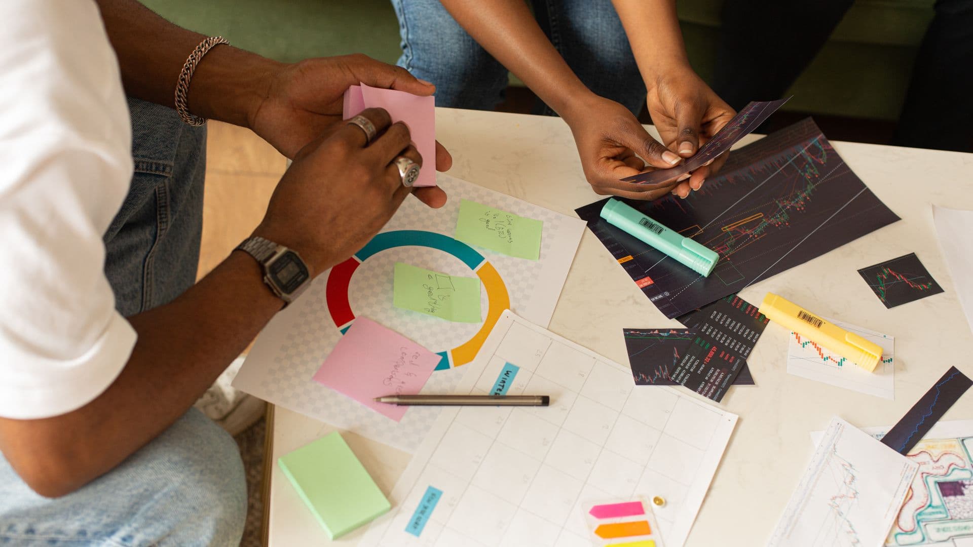 Hands arrange printed charts, sticky notes, and markers on a planning table.